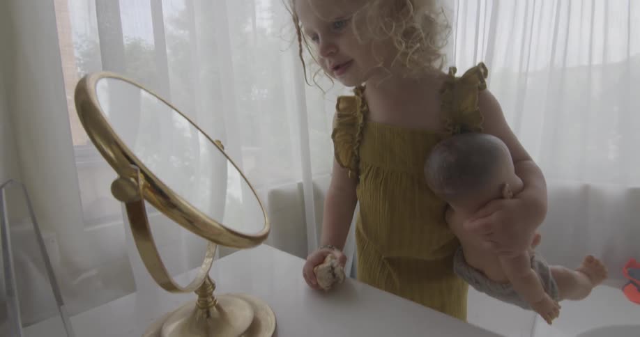 Little girl holds her doll as she looks into a round mirror