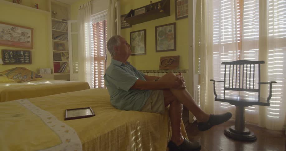 An elderly man is sitting on the edge of a bed with a yellow comforter looking out the window in a room painted yellow with a picture in a frame beside him