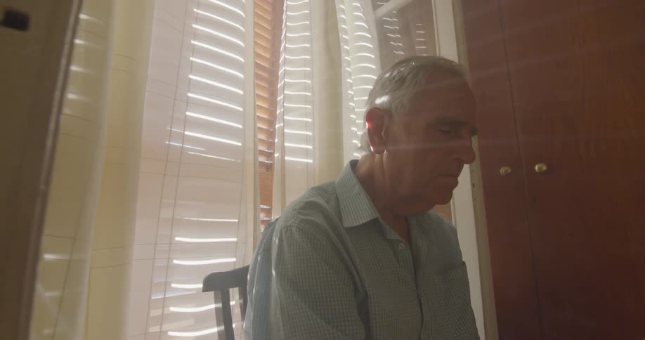 Old man sits on the chair in front of the window and looks down in a room