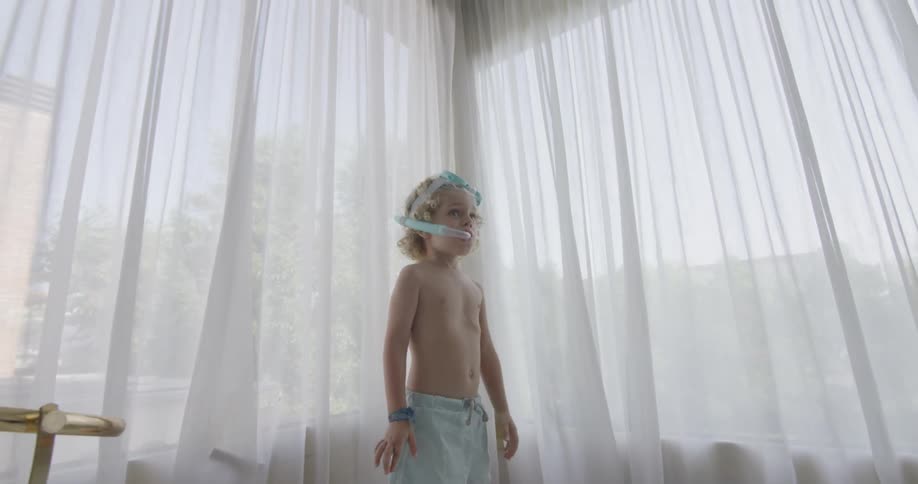 A young boy stands next to a wide curtain while he wears swimming trunks and a snorkel