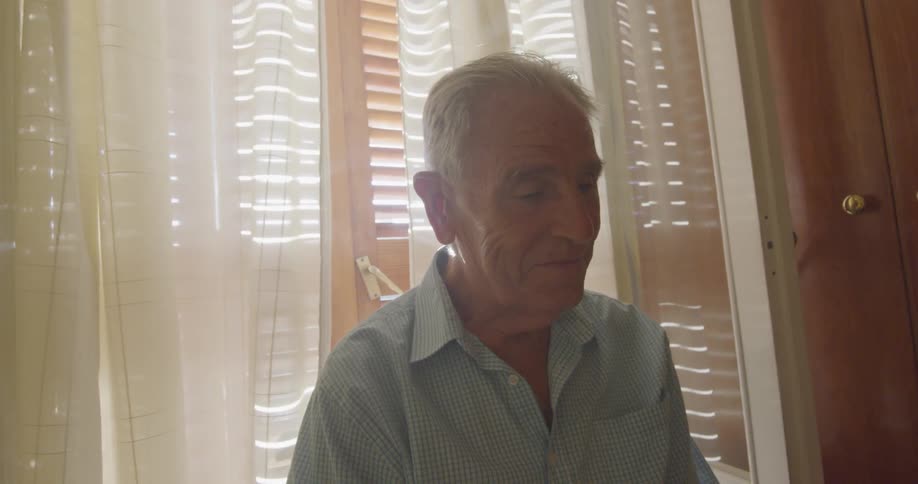 Senior man sitting on the chair in front of the window talks to someone in the room