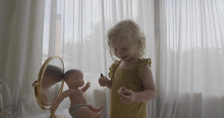 A little girl holds a tube of lipstick while she stands in front of a doll resting on a small mirror