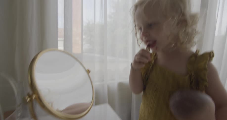 A young blonde girl smiles while putting lipstick on her face