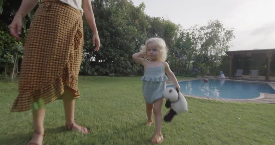 Two blonde children carry their stuffed animals as they run and walk past their mom in the back yard