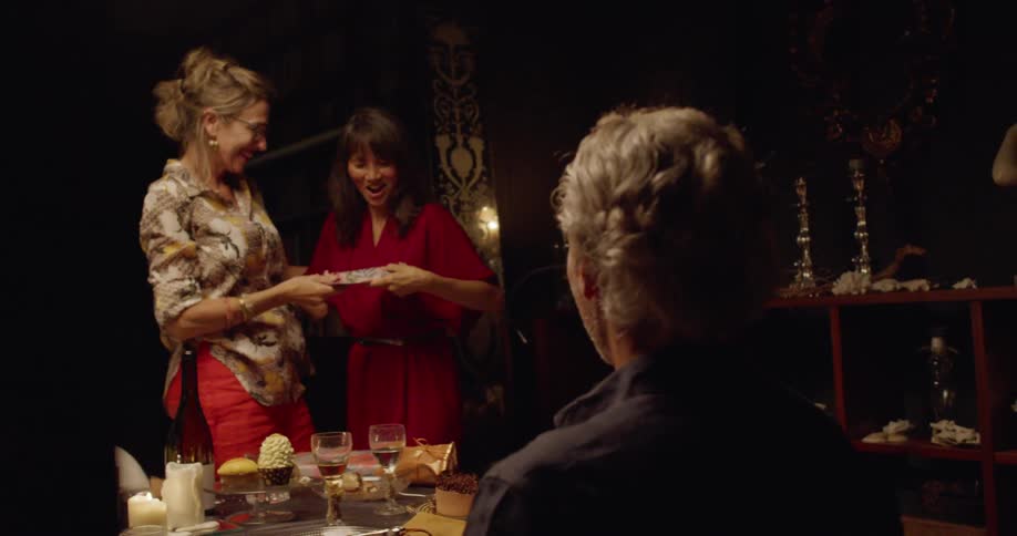 Two women hug and exchange something at a dinner table