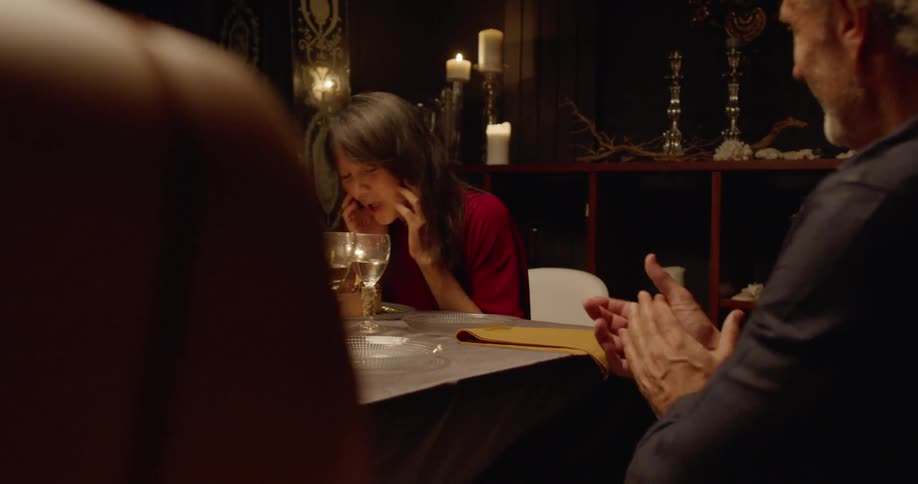 Friends sit around dining table clapping as birthday girl blows candles on cake out