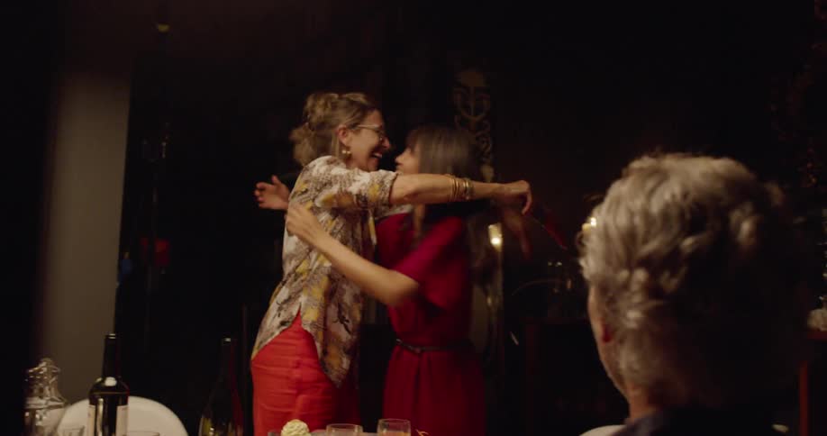 Two women stand up from the dinner table and hug each other