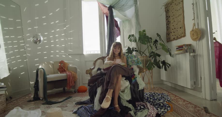 A young girl is sitting in a chair with clothes tossed all over the furniture and floor reading a book while a disco ball reflects on the walls near the back corner of the room