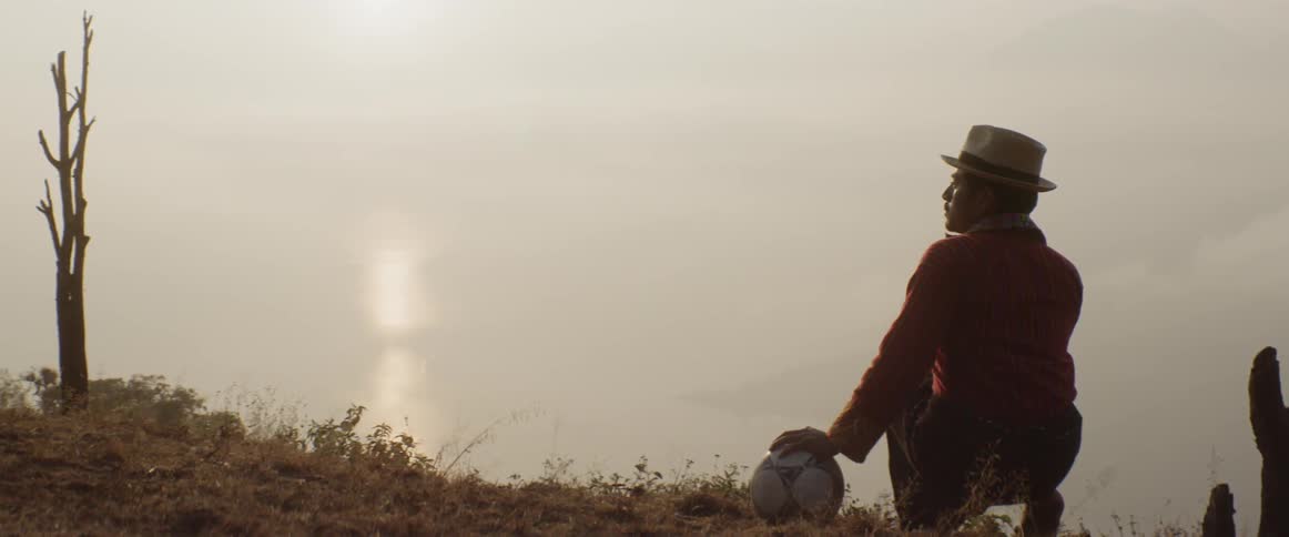 Man crouches outdoors on shore with hand on soccer ball