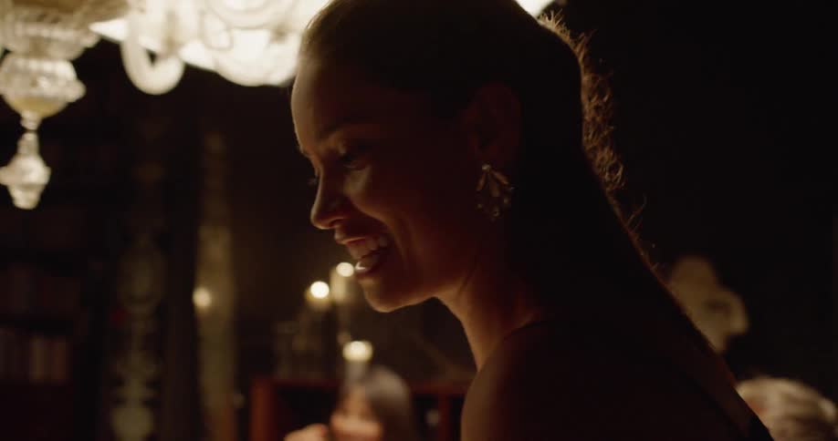 A woman in a dress stands in room lit by a large chandelier as she speaks with other people in the room