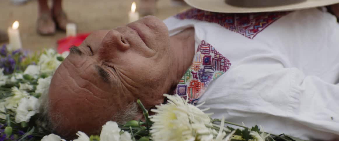 An old man lies on a bed of flowers grimacing and surrounded by candles
