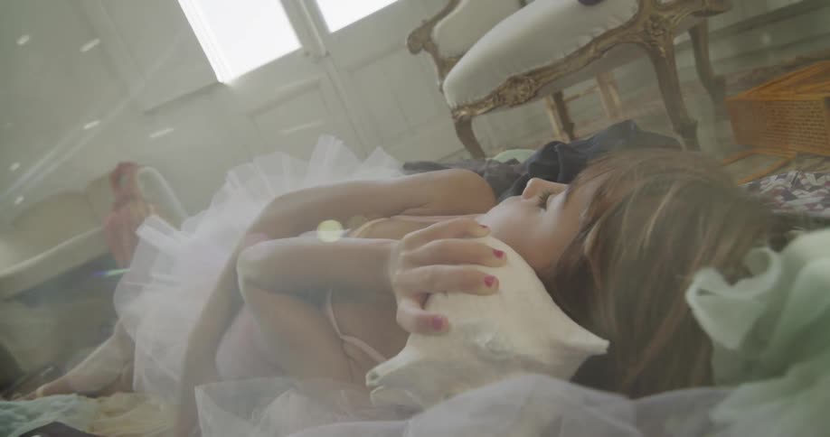 Young girl in a ballerina outfit listens to a seashell as she lays on the bed