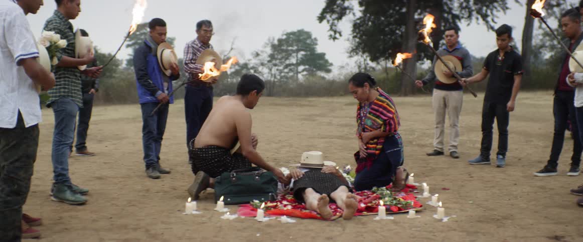 A man lies on the grass while surrounded by candles and flowers as other people surround him as part of a ritual
