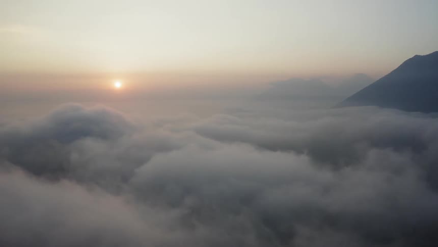 High above a thick layer of fluffy clouds a mountain stands tall as the sun sits low in the sky beyond it