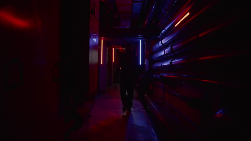 A person walking through a narrow and dimly lit hallway lined with pipes