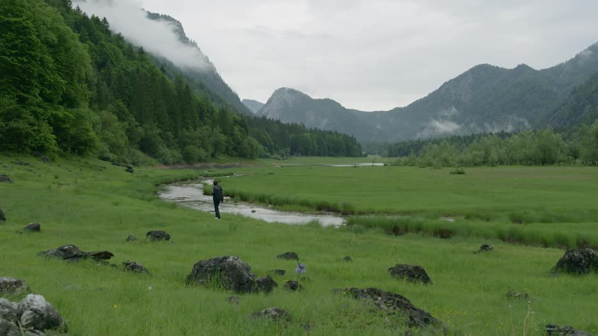A man is wearing a backpack hiking along a stream running through a field surrounded in forest and mountains