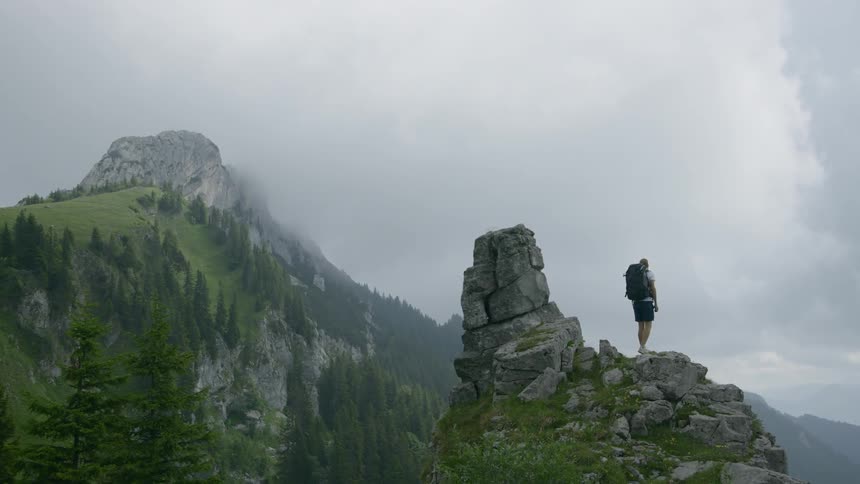 A male hike with a large backpack ascents a rocky prominence and looks out up a tall mountain in the distance