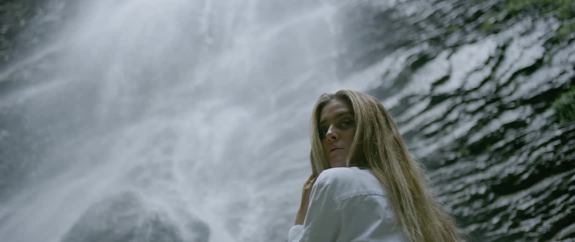 A woman stands in front of a waterfall then turns around to look behind her
