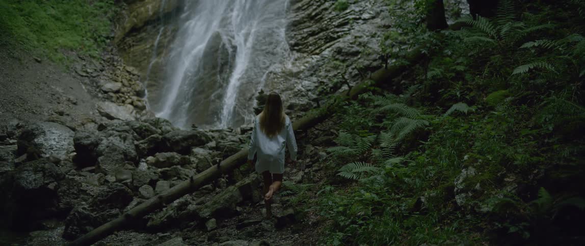 A woman in a large white shirt walking barefoot towards a waterfall