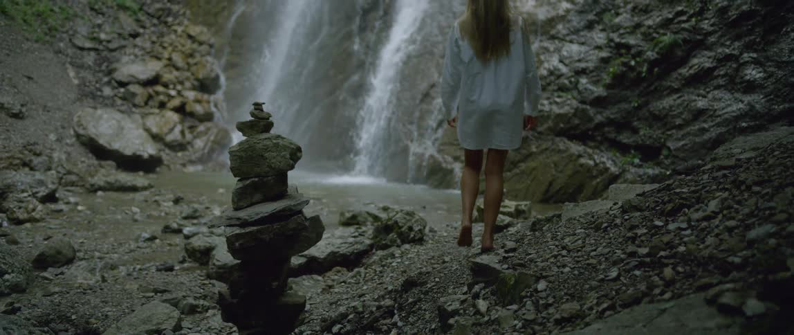 A woman wearing only a linen shirt walks towards a small waterfall