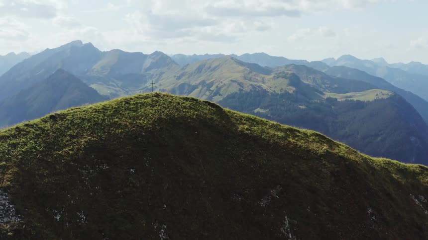 A cross sits on top of a greenery covered mountain under a cloudy sky