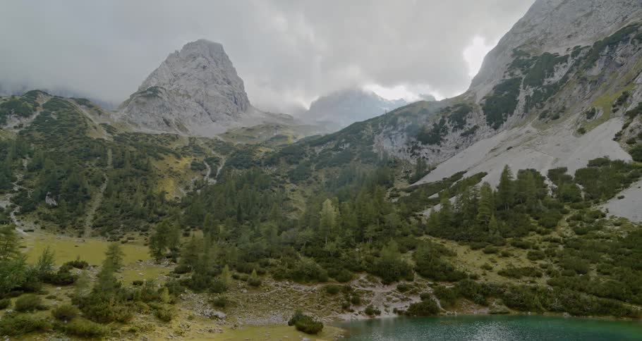 Underneath a dense layer of clouds a mountain range is bordered by thin forests and a lake