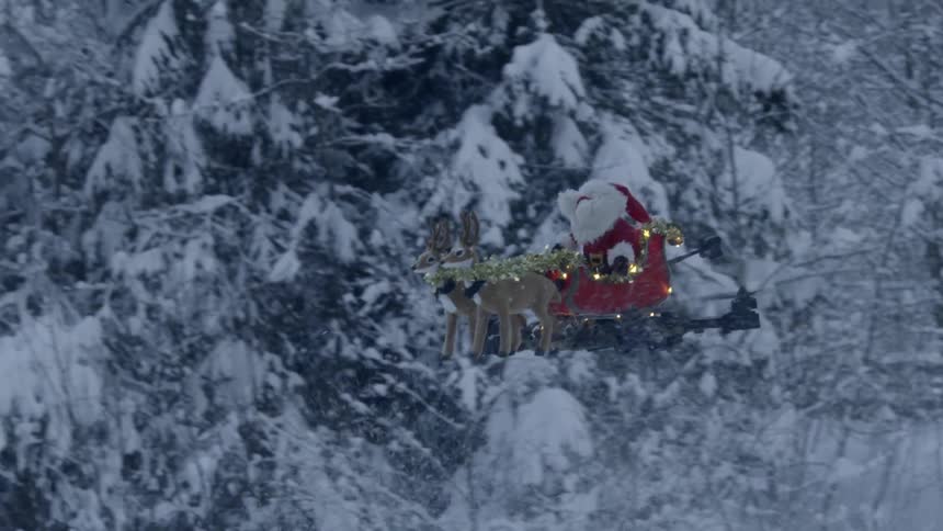 A flying drone is decorated with toy reindeer pulling santa claus in a sleigh as it hovers through a snow covered forest
