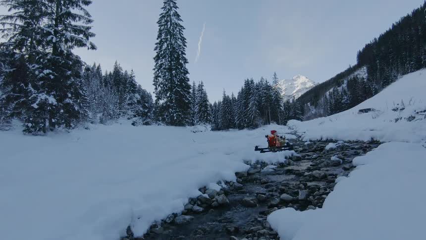 A remotely controlled drone with santa claus and reindeer figures on top flying over a rocky stream in a snowy countryside
