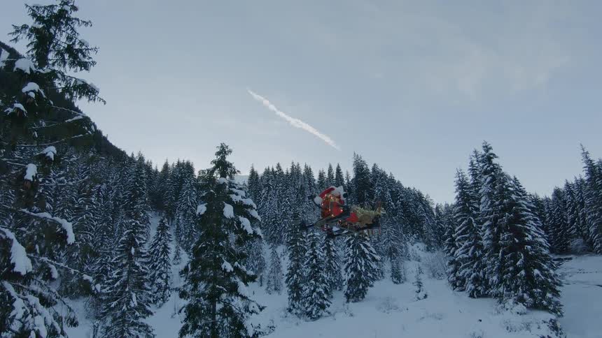 A small flying drone is decorated with a santa and reindeer figurines as it flies above a snow covered forest