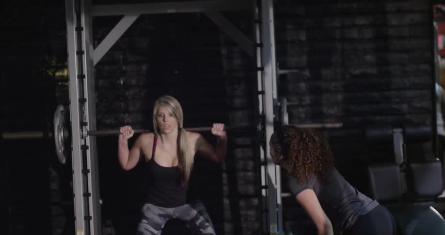 Woman squatting with weights in a gym while another woman encourages her