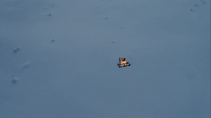 A small quad copter drone with many lights on it flies over a snow covered outdoor landscape