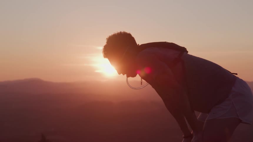A man is bent over with hands on knees catching his breath while the sun sets