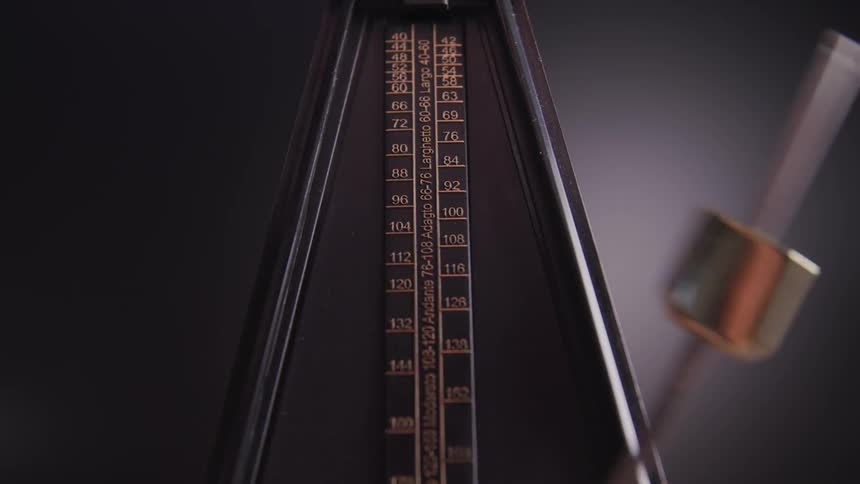 An old fashioned metronome swings back and forth in front of a gray background