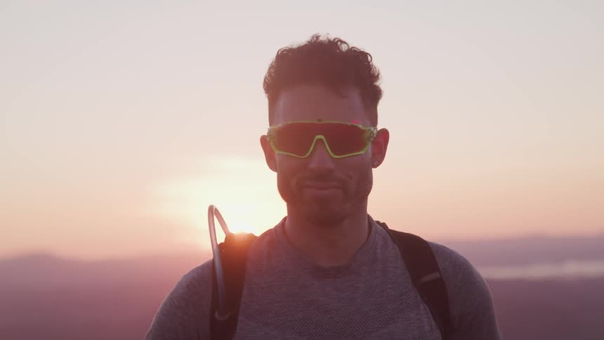 A man wearing a hydration backpack and sunglasses stands looking straight ahead at the camera as the sun sets on the horizon behind him