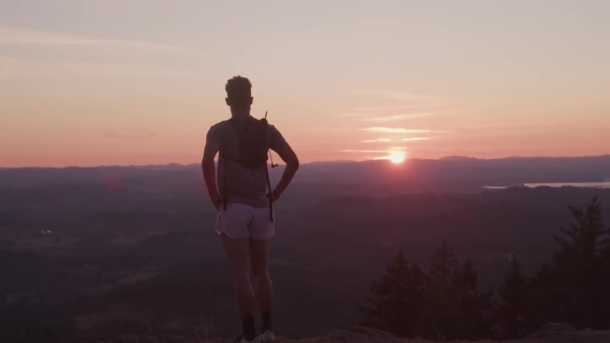 A hiker in shorts standing on ledge looking out over the countryside below at sunset