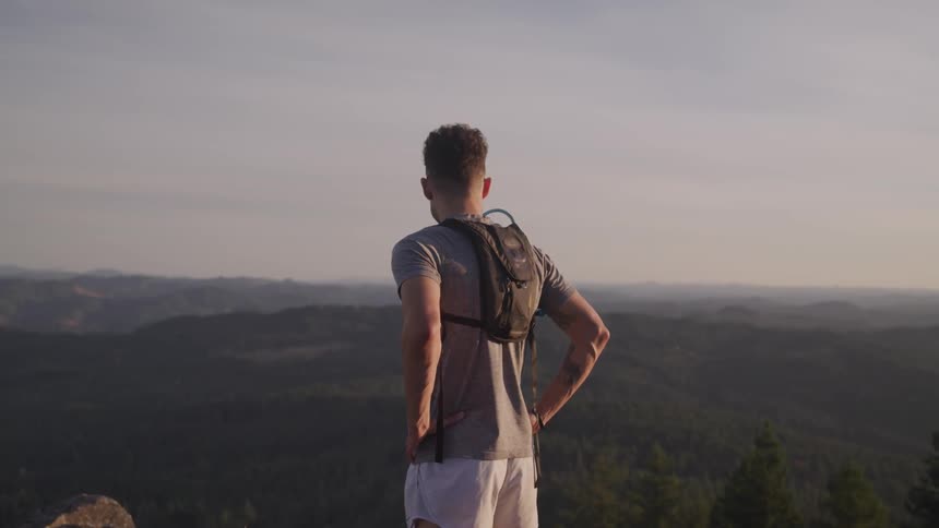 A man stands at the top of a mountain overlooking a forested valley