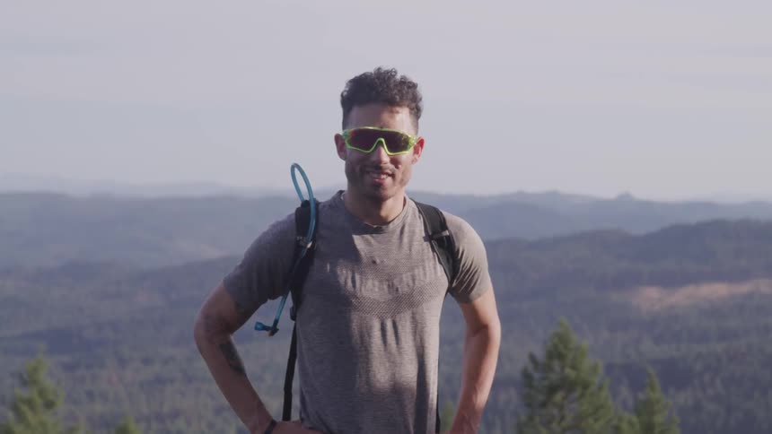 A man stands atop a mountain with a backpack and sunglasses