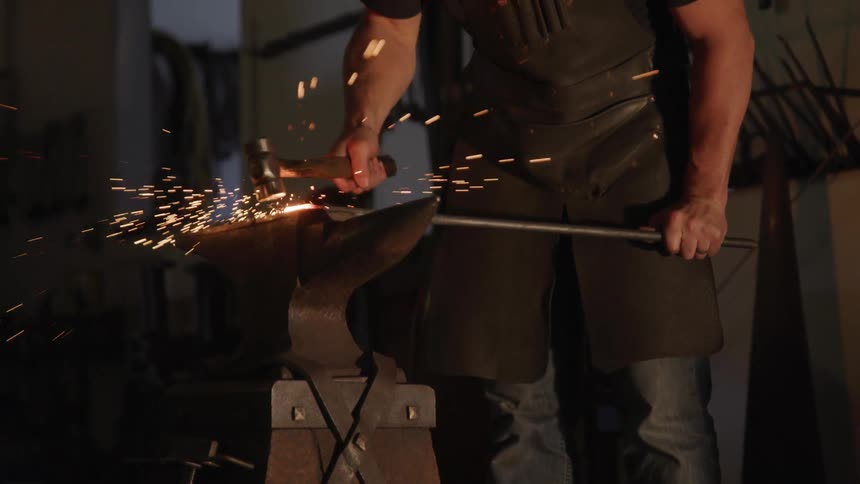 A man holds a metal rod atop and anvil and strikes the glowing tip of it with a blacksmith hammer