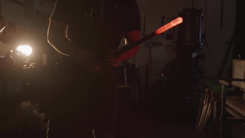 A metal worker withdraws a glowing hot piece of metal from a furnace and prepares to set it down on a work surface