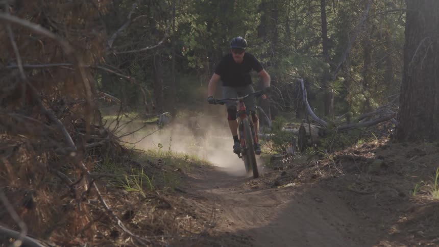 A mountain bike rider comes around a corner and makes a jump on a dirt trail in the woods