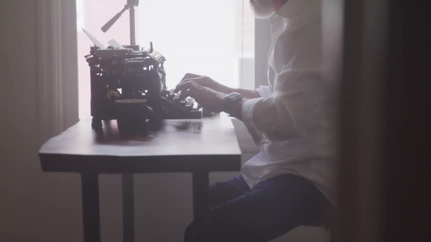 Man sitting at a table next to a window typing on a vintage typewriter