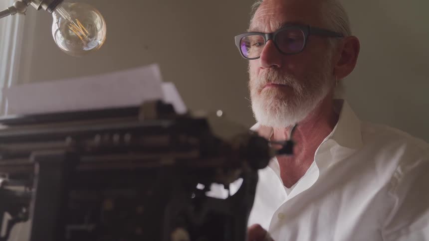 Old bearded man types on a typewriter in a room