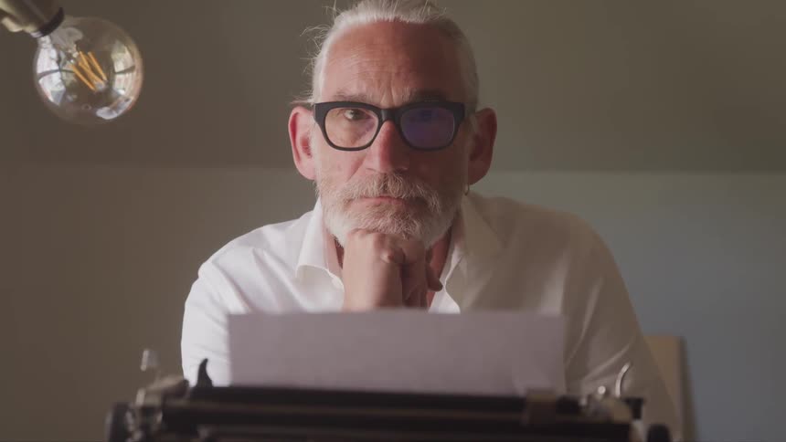 An older man with white hair and blackrimmed eyeglasses is sitting in front of a typewriter and looks up to stare at the camera