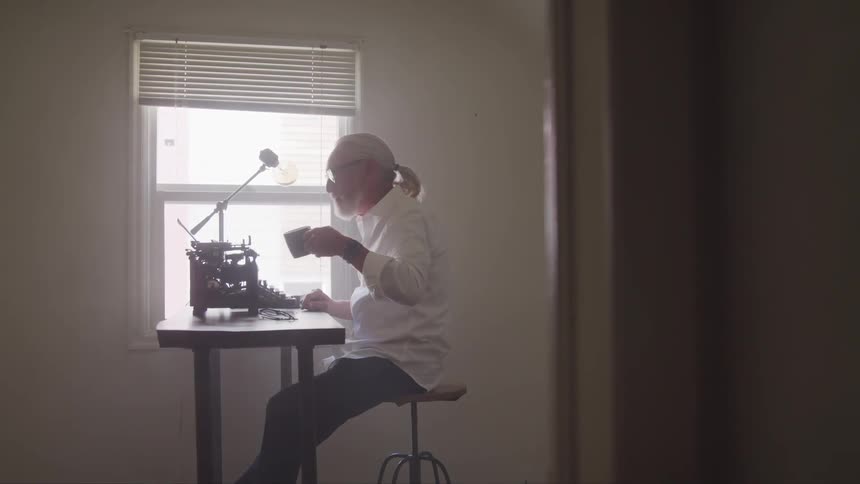 A man takes a drink from a mug while sitting in front of a vintage typewriter