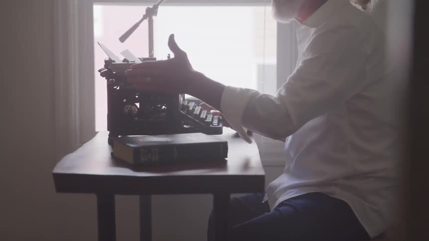 Senior man sittin at the table types on the typewriter next to the window in a room