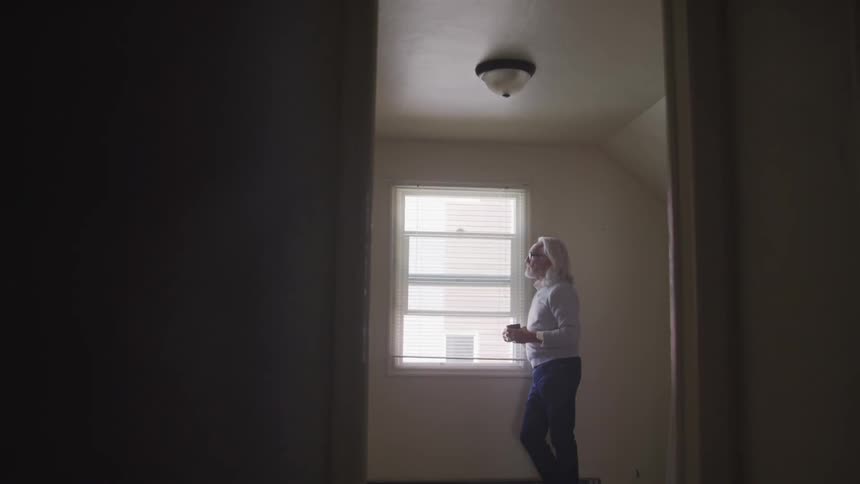 Senior grey long hair man stands next to the window and looks out in a room during the day