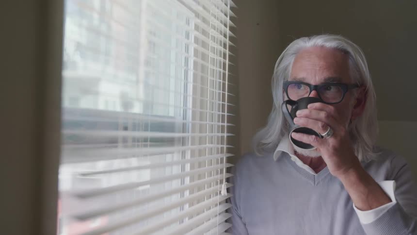 Elderly white male looking out a window while drinking a cup of coffee