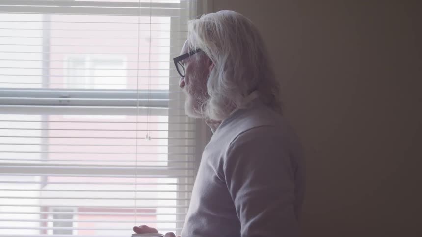 Long grey hair man holding a cup stands by the window and looks out in a room
