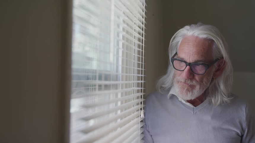 Senior long hair man stands by the window and looks out in a room