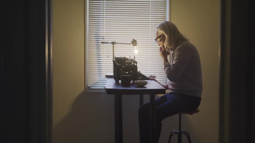A an older white male is sitting on a stool at a small desk in front of a vintage typewriter and is smoking something using his left hand